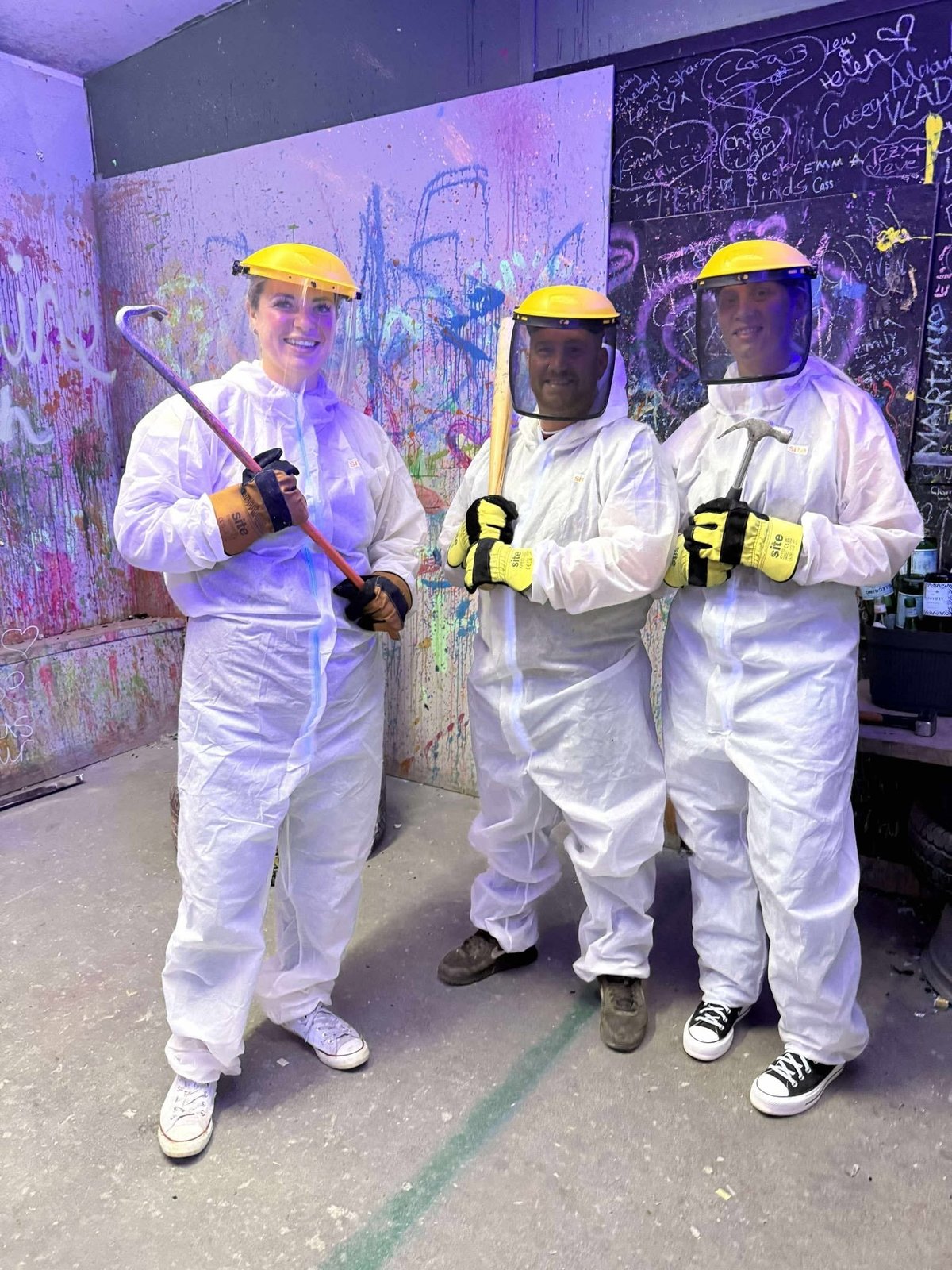 Three people in white protective suits, yellow hard hats, and face shields pose together in front of a colorful graffiti wall holding tools